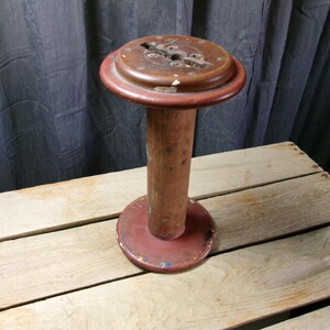 May include: A vintage wooden industrial spool featuring a cylindrical central shaft and two circular end plates. The wood displays a weathered finish with remnants of red paint and scattered paint splatters across the surface of the spool.