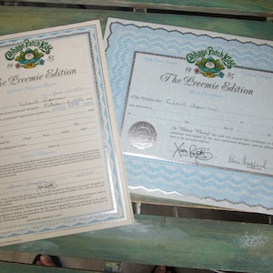 May include: Two Cabbage Patch Kids Preemie Edition birth certificates. The certificates are printed on white paper with blue and silver accents. The text on the certificates reads "Cabbage Patch Kids 1985 The Preemie Edition".