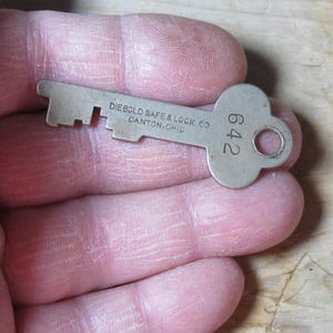 May include: A vintage metal key with the text "DIEBOLD SAFE & LOCK CO CANTON, OHIO" and the number "642". The key is silver-colored and has a unique shape with a circular hole at the end. It is held in a person's hand.