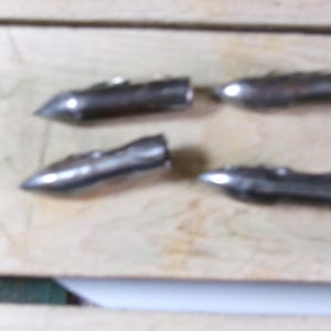 May include: Four small, metallic, bullet-shaped objects with pointed tips and ridged bodies are arranged on a light-colored wooden surface. The objects appear to be made of a silver-colored metal.