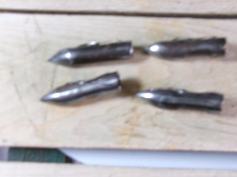 May include: Four small, metallic, bullet-shaped objects with pointed tips and ridged bodies are arranged on a light-colored wooden surface. The objects appear to be made of a silver-colored metal.