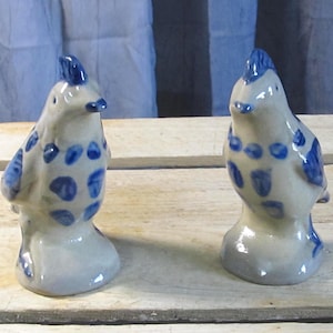 May include: Two ceramic bird figurines. The birds are white with blue accents, including spots and crests. The figurines have a rounded shape and are standing upright. The background is a light blue and white.