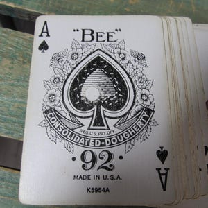 Puede incluir: Una baraja de cartas de juego vintage, con el As de Picas. El dise&ntilde;o de la carta incluye un gran s&iacute;mbolo de picas con acentos florales, la palabra "BEE" y el texto "CONSOLIDATED-DOUGHERTY". Las cartas est&aacute;n marcadas con "92" y "MADE IN U.S.A."