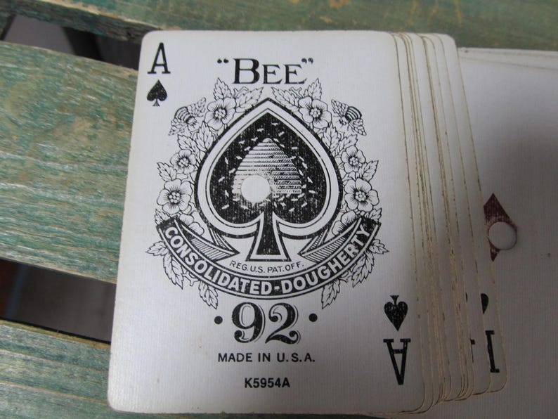 Puede incluir: Una baraja de cartas de juego vintage, con el As de Picas. El dise&ntilde;o de la carta incluye un gran s&iacute;mbolo de picas con acentos florales, la palabra "BEE" y el texto "CONSOLIDATED-DOUGHERTY". Las cartas est&aacute;n marcadas con "92" y "MADE IN U.S.A."