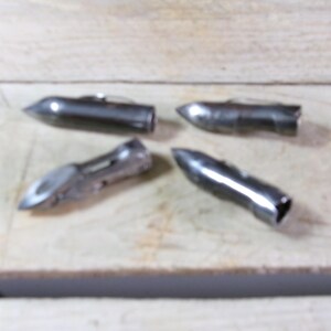 May include: Four metallic silver bullet-shaped objects with pointed tips and open ends are arranged on a light-colored wooden surface. The objects appear to be made of metal and have a shiny finish.
