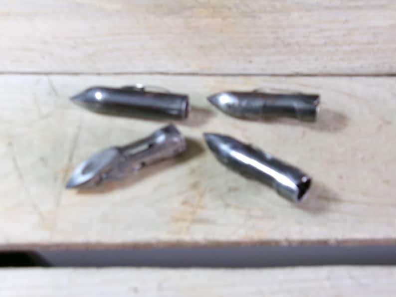 May include: Four metallic silver bullet-shaped objects with pointed tips and open ends are arranged on a light-colored wooden surface. The objects appear to be made of metal and have a shiny finish.