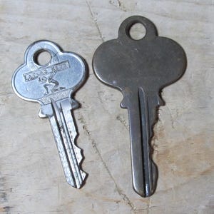 May include: Two vintage keys rest on a wooden surface. One key is silver-toned with the text "ATLAS KEYS" and a graphic of a person. The other key is a darker, aged metal. Both keys have a loop at the top.