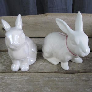 May include: Two white ceramic bunny figurines. One bunny is sitting with its ears up and the other bunny is sitting with its ears down. Both bunnies have a glossy finish.