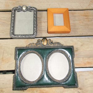 May include: Three decorative picture frames. A silver rectangular frame with a bow detail, a small wooden frame, and a green and gold double oval frame. The frames are empty, ready for photos.