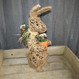 May include: A brown twine bunny rabbit holding a small terracotta pot with white flowers. The bunny is standing on a wooden surface.