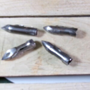 May include: Four silver-colored metal bullet-shaped objects with pointed tips. The objects are arranged on a light-colored wooden surface. The objects appear to be made of a shiny, metallic material.