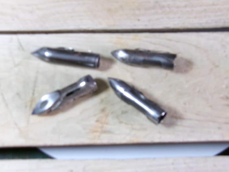 May include: Four silver-colored metal bullet-shaped objects with pointed tips. The objects are arranged on a light-colored wooden surface. The objects appear to be made of a shiny, metallic material.