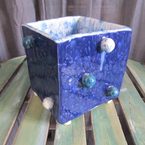 May include: Square blue ceramic planter with white and blue glaze. The planter has six round white and blue accents on the sides and bottom.