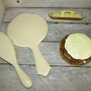May include: A vintage vanity set with a hand mirror, a brush, and a round, amber-colored container with a yellow lid. The set is made of plastic and has a cream color.