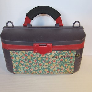 May include: A purple and red vintage cosmetic case with a floral pattern and a black handle. The case has a small metal latch and a "QC" logo.