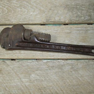 Op de afbeelding: Een vintage, roestige Ridgid pijptang, liggend op een houten oppervlak. De sleutel is donkerbruin met zichtbare tekst die "TRADE RIDGID MARK" en "10" luidt. De tool heeft een getande bek en een verstelbaar mechanisme.