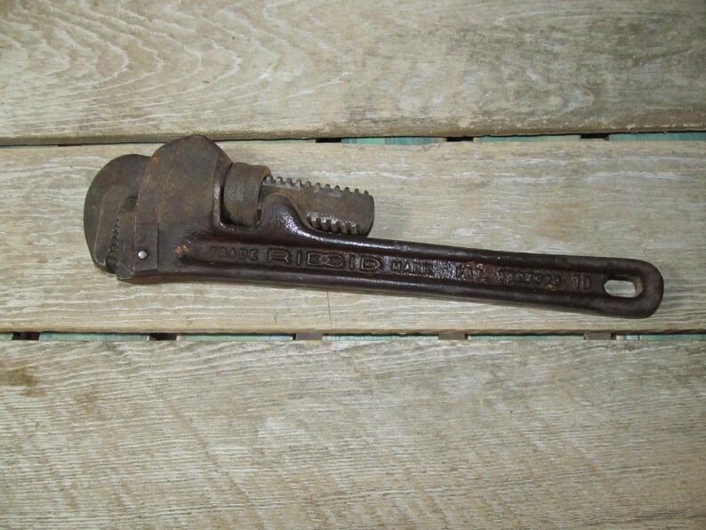 Op de afbeelding: Een vintage, roestige Ridgid pijptang, liggend op een houten oppervlak. De sleutel is donkerbruin met zichtbare tekst die "TRADE RIDGID MARK" en "10" luidt. De tool heeft een getande bek en een verstelbaar mechanisme.