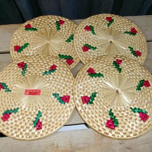 May include: Four round woven coasters made of light tan natural fibers, each featuring a repeating pattern of small red and green floral accents. One coaster includes a small rectangular red label with white text in the center.