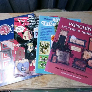 May include: A collection of four craft instruction booklets featuring colorful covers. The books include titles such as The Ornament, McCall's Jiffy Tree, and Punchin' Letters & Numbers, displaying various patterns for quilts, ornaments, and lettering projects.
