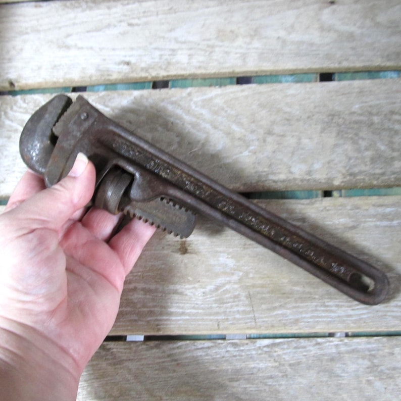 Op de afbeelding: Een vintage, roestige pijpsleutel. De sleutel heeft een donkerbruine kleur en heeft een lange handgreep met een getande bek. De verstelbare bek is open. De sleutel wordt in een hand gehouden.