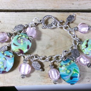 May include: A silver-toned charm bracelet featuring a mix of round, translucent light pink glass beads and larger, oval-shaped glass beads with swirling patterns of teal, green, and purple, accented by small silver metal leaf-shaped charms.