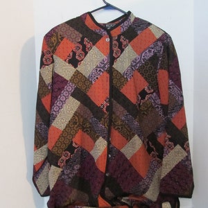May include: A long-sleeved button-down shirt with a patchwork design. The shirt features a variety of geometric shapes in shades of orange, purple, brown, and black. The collar and button placket are trimmed in black.