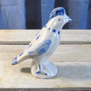 May include: A white ceramic bird figurine with blue accents. The bird has a rounded body, a small beak, and blue spots and wing details. The figurine is standing on a small, round base. The bird is a decorative item.