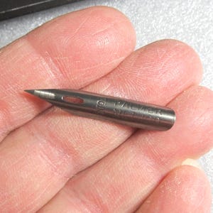 May include: A close-up of a silver-colored pen nib with the text "O.M. PALMER METHOD" engraved on the side. The nib has a sharp, pointed tip and a small oval-shaped hole near the base.