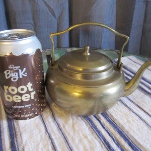 May include: A brass teapot with a lid and a handle. The teapot is sitting on a white and blue striped tablecloth next to a can of Kroger Big K Root Beer.
