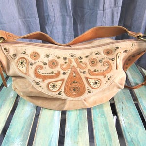May include: A tan suede shoulder bag with brown leather straps and accents. The bag features decorative stitching and metal studs. The front of the bag has an ornate design with paisley and circular patterns.