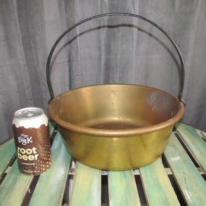 May include: A large, round, brass bowl with a black metal handle. The bowl is sitting on a wooden surface.