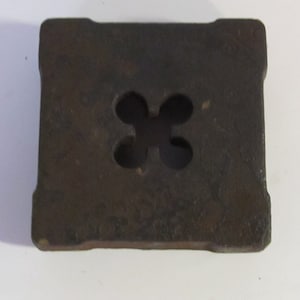 May include: A square, dark brown metal object with a four-leaf clover-shaped hole in the center. The edges are slightly rounded. The object appears to be made of cast iron and has a rough, textured surface.