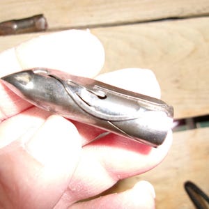 May include: A silver-colored, bullet-shaped object with a pointed tip. The object has a curved design on its side and appears to be made of metal. Another similar object is visible in the background.