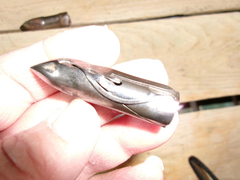 May include: A silver-colored, bullet-shaped object with a pointed tip. The object has a curved design on its side and appears to be made of metal. Another similar object is visible in the background.