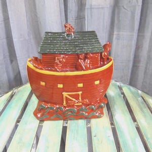 May include: A red and green ceramic cookie jar shaped like Noah's Ark. The ark has a green roof and a brown door. There are figures of people and animals on the ark.