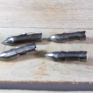 May include: Four small, metallic silver objects with a bullet-like shape are arranged on a light-colored wooden surface. Each object has a pointed end and a wider, cylindrical body. The objects appear to be made of metal.
