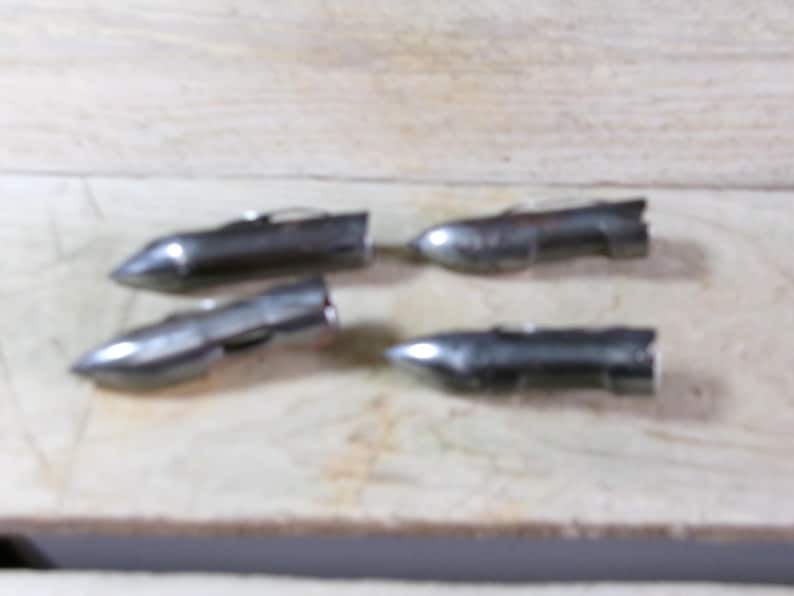May include: Four small, metallic silver objects with a bullet-like shape are arranged on a light-colored wooden surface. Each object has a pointed end and a wider, cylindrical body. The objects appear to be made of metal.