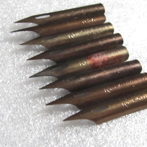 May include: A collection of vintage calligraphy pen nibs. The nibs are made of bronze-colored metal and have sharp, pointed tips. Some nibs have the text "USA" and "2 LAURA INC" engraved on them. The nibs are arranged diagonally on a white surface.