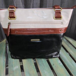 May include: A black, brown, and white leather tote bag with a zipper closure and a gold metal logo that reads "Fille de Grace".