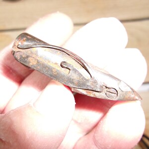 May include: A small, antique metal tool with a curved design and a pointed end. The tool has a rusty, aged appearance, with visible wear and tear. It features a keyhole-shaped opening and a small screw. The object is held in a hand.