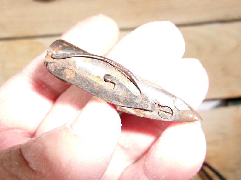 May include: A small, antique metal tool with a curved design and a pointed end. The tool has a rusty, aged appearance, with visible wear and tear. It features a keyhole-shaped opening and a small screw. The object is held in a hand.