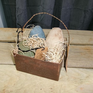 May include: A rustic brown metal basket with a woven handle, filled with decorative items. The basket contains a blue stone, a beige stone, and a green stone, nestled in straw. A small tag is attached.