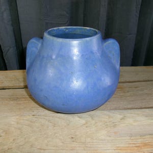 May include: A round, blue ceramic vase with a smooth, matte finish and two integrated side handles. The vessel features a wide, open circular mouth and a bulbous body, showcasing a consistent, solid blue color throughout its entire exterior surface.