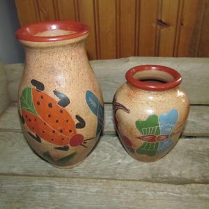 May include: Two terracotta vases with hand-painted designs. The larger vase features a ladybug with orange and black spots, green leaves, and blue accents. The smaller vase has a floral design with green, blue, and red accents.