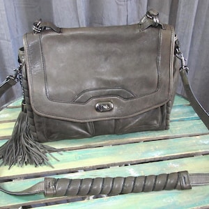 May include: A gray leather crossbody bag with a flap closure and a long adjustable strap. The bag features a decorative tassel and a braided handle detail. The bag is sitting on a multi-colored wooden surface.