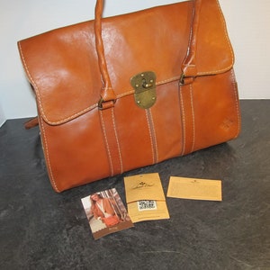 May include: A brown leather satchel bag with a brass lock and buckle. The bag has a top handle and a shoulder strap. The bag is sitting on a dark surface with two small cards.