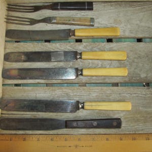 May include: A set of six vintage kitchen utensils, including a fork, a butter knife, and four table knives. The utensils have wooden handles and metal blades. The utensils are arranged on a wooden surface with a ruler.