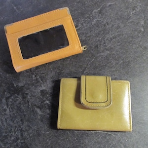May include: Two leather wallets. One is a light brown cardholder with a clear window. The other is a light green bi-fold wallet with a flap closure. Both wallets are on a dark gray surface.
