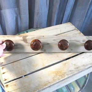 May include: A rustic wooden coat rack with four dark brown, glossy knobs. The knobs are evenly spaced along the length of the light-colored, weathered wood. The rack is designed for hanging items and adds a touch of natural decor.