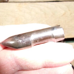 May include: A metallic bullet with a pointed tip and a cylindrical body. The bullet has a silver finish with some darker spots, suggesting age or use. The bullet is held in a hand.